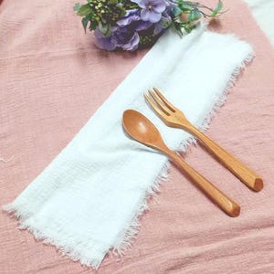 Könnte beinhalten: Ein Holzlöffel und eine Gabel liegen auf einer weißen Serviette mit Fransen, die auf einem rosa Stoff liegt. Ein kleiner Strauß lila Blumen liegt über der Serviette. Der Löffel und die Gabel sind aus hellem Holz.