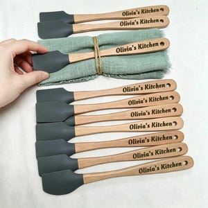 Könnte beinhalten: Eine Sammlung von Silikonspateln mit hellen Holzgriffen, die jeweils mit dem Text Olivia's Kitchen graviert sind. Die Spatel haben dunkelgraue Silikonköpfe und sind neben einer gefalteten, mit Bindfaden umwickelten salbeigrünen Serviette.