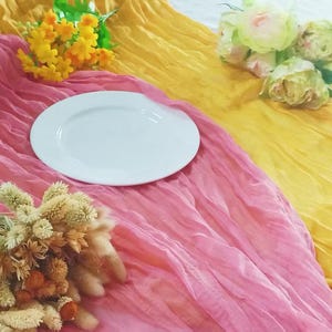 Könnte beinhalten: Ein weißer Teller liegt auf einer rosa und gelben, durchsichtigen Stofftischdecke. Der Stoff ist gerafft und gekräuselt, was einen weichen und eleganten Look erzeugt.