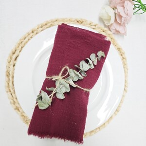 May include: A folded claret napkin, decorated with a sprig of eucalyptus and twine, sits on a white plate. The plate is on a woven placemat, with a bouquet of pink and white flowers in the background. The image presents a rustic, elegant table setting.
