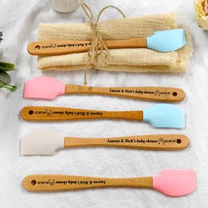 Könnte beinhalten: Fünf kleine Silikonspatel mit Holzgriffen, jeweils graviert mit dem Text Lauren & Nick's baby shower und dem Datum 08.09.25. Die Spatel haben hellblaue, rosa und weiße Köpfe und sind horizontal auf einem gefalteten beigen Tuch angeordnet.