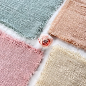 Op de afbeelding: Vier vierkante stukken stof in verschillende kleuren: lichtblauw, perzik, roze en beige. De stof heeft een textuur, geweven look en rafelige randen. Een roze bloem bevindt zich in het midden van de vierkante stukken stof.