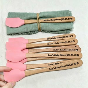 Könnte beinhalten: Ein Set aus hellrosa Silikonspateln mit Holzgriffen, die jeweils mit einem Namen und Datum für eine Babyparty graviert sind. Die Spatel liegen auf einer salbeigrünen Stoffserviette, die mit Bindfaden zusammengebunden ist, mit Text wie Marie's Baby Shower 08.08.25.