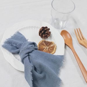 Può includere: Un piatto bianco con un tovagliolo blu annodato, un cucchiaio e una forchetta di legno, un bicchiere trasparente, una pigna e una fetta di arancia secca. Il tovagliolo ha un bordo sfilacciato. Gli oggetti sono disposti su una superficie bianca.