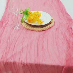 May include: A pink crinkled table runner with a white plate, a woven placemat, and a glass of water. Yellow flowers are arranged on the plate.