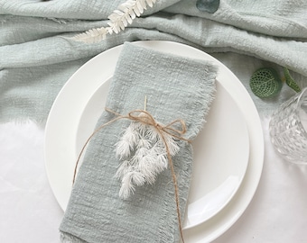 May include: A light green linen napkin with frayed edges is folded and tied with twine. A sprig of dried white flowers is tied to the twine. The napkin is placed on a white plate.