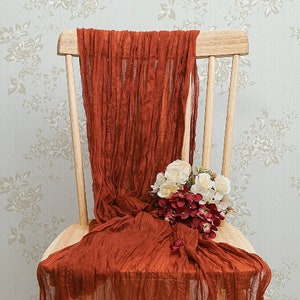 Puede incluir: Una silla de madera con una tela de color óxido drapeada sobre ella. La tela está recogida y arrugada en el asiento y el respaldo de la silla. Un ramo de flores blancas y rojas se coloca sobre la tela.