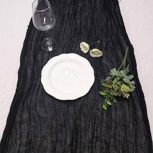 May include: A black crinkled fabric table runner with a white plate, a wine glass, and floral accents. The plate has a decorative edge. The runner is draped over a white surface, creating a contrast.