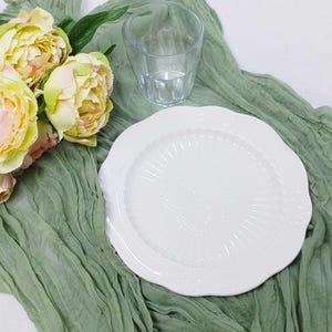 May include: A white plate with a scalloped edge and floral detailing rests on sage green fabric. A clear glass and a bouquet of pink and yellow peonies are also present, creating a soft, elegant aesthetic.