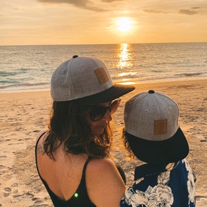 Könnte beinhalten: Eine Frau und ein Kind tragen passende graue und schwarze Baseballkappen mit einem braunen Lederpatch auf der Vorderseite. Die Frau trägt ein schwarzes Tanktop und das Kind trägt ein blau-weißes Blumenhemd. Sie stehen an einem Sandstrand mit dem Meer im Hintergrund.