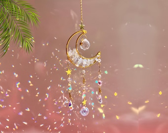 May include: A gold crescent moon-shaped wind chime with crystal beads and stars hanging from it. The wind chime is suspended from a gold chain.