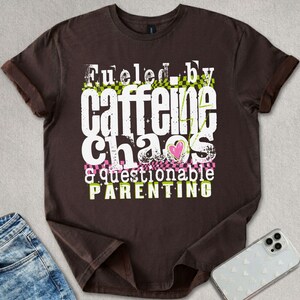 May include: A brown t-shirt featuring the text "Fueled by Caffeine Chaos & Questionable Parenting" in white, green, and pink. The design includes a heart graphic and a checkered pattern.