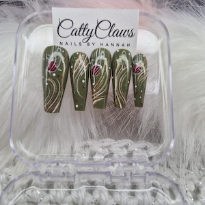 May include: Olive green press-on nails with a floral design in a clear plastic case. The nails feature gold and white swirls, red accents, and small white dots. The text "Catty Claws NAILS BY HANNAH" is visible on a white card.