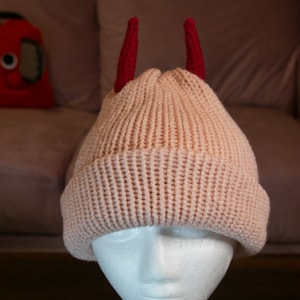Könnte beinhalten: Eine rosafarbene Strickmütze mit roten Hörnern. Die Mütze sitzt auf einem weißen Styropor-Kopf.