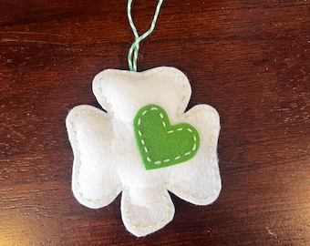 May include: A white felt shamrock ornament with a green felt heart in the center. The ornament has a green and white twine hanger.