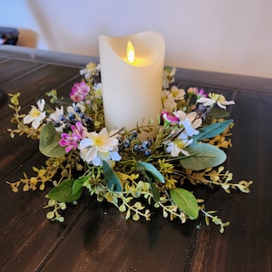 May include: A white pillar candle with a flickering flame sits in the center of a floral wreath. The wreath is made of green leaves, white flowers, and small purple flowers.