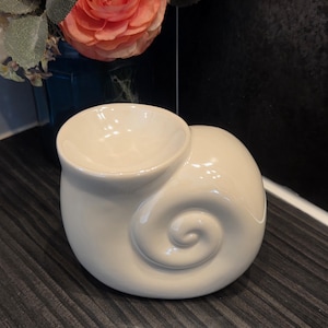 May include: A cream-coloured ceramic seashell-shaped bowl. The bowl has a rounded spiral design and a small, circular opening at the top. The bowl is on a dark, textured surface.