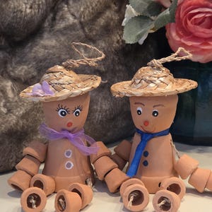 May include: Two terracotta pot figures, each wearing a straw hat and painted faces. One has a purple ribbon, the other a blue tie. The figures have pot segments for limbs, and are sitting on a white surface.