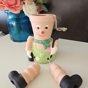May include: A ceramic planter shaped like a person with a green apron, brown legs, and black shoes. The planter has a smiling face with a red nose and a green scarf.