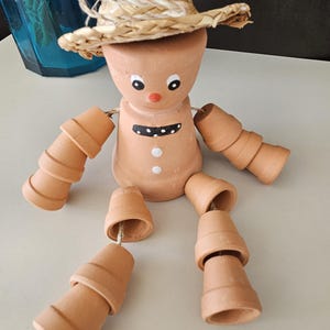 May include: A whimsical terracotta figurine of a person wearing a straw hat and painted with a smiling face. The figurine's arms and legs are made of stacked terracotta pots.