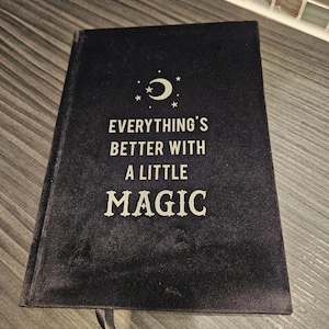 May include: A black velvet journal with a silver crescent moon and stars design. The text on the cover reads "Everything's better with a little magic."