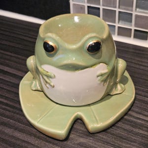 May include: A green ceramic frog egg cup with black eyes and a gold rim sits on a green ceramic lily pad.
