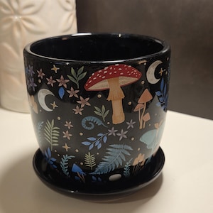 May include: Black ceramic plant pot with a matching saucer. The pot is decorated with a whimsical design featuring mushrooms, crescent moons, and various floral and foliage elements in shades of red, blue, and green.