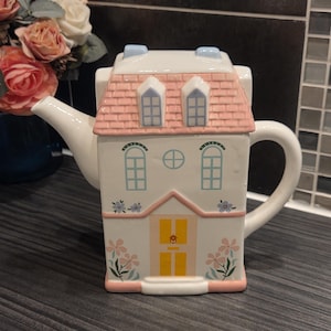 May include: A house-shaped ceramic teapot in white, with a pink roof and blue window details. The design includes a yellow door and floral patterns. The teapot's handle is curved. A decorative and unique item for tea lovers.