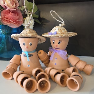 May include: Two terracotta pot figures with straw hats and rope hangers. One figure has a blue scarf and the other a purple bow. Several small terracotta pots are scattered in front of them. The figures have painted eyes and button details.