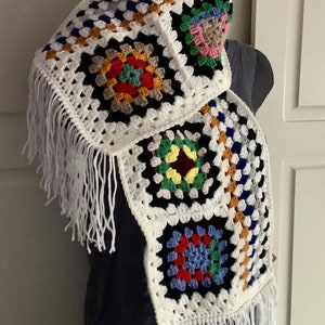 Peut inclure: Une écharpe en crochet blanche avec des carrés de grand-mère colorés et des franges blanches. L'écharpe est faite avec une variété de couleurs, dont le rouge, le bleu, le vert, le jaune et le noir.