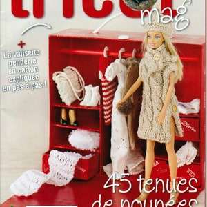 Peut inclure: Une armoire rouge de style maison de poupée avec une poupée portant un pull en tricot beige et un chapeau blanc. L'armoire a des étagères avec divers articles en tricot, dont une écharpe en tricot blanc, un chapeau en tricot blanc et un sac en tricot blanc. L'armoire a également un tiroir avec une écharpe en tricot blanc. Le texte sur la couverture du magazine est "45 tenues de poupées mannequins".