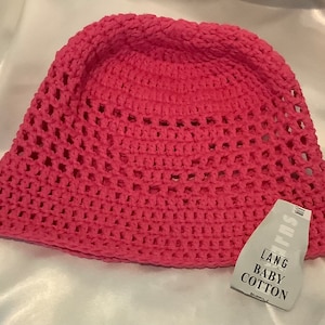 Peut inclure: Chapeau seau en crochet rose avec une étiquette blanche indiquant "LANG BABY COTTON" et "BAUMWOLLE".