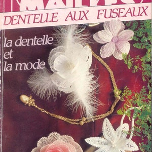 Peut inclure: Une couverture de livre vintage avec un fond rose et le titre "Mailles Dentelle Aux Fuseaux" en texte blanc. La couverture présente également une plume blanche, un collier de chaîne dorée et trois fleurs en dentelle blanche.