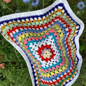 Peut inclure: Une couverture en crochet colorée avec une bordure blanche et une bordure intérieure bleue et blanche. La couverture est faite de carrés de grand-mère dans une variété de couleurs, y compris le rouge, l'orange, le jaune, le vert, le bleu, le violet et le rose.