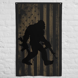 May include: A black silhouette of Bigfoot holding two rifles, standing in front of an American flag with a distressed, vintage look.