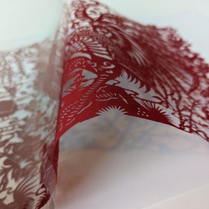 May include: Red paper cut-out art with intricate designs of birds and flowers. The paper is folded over, revealing the delicate details of the cut-out.