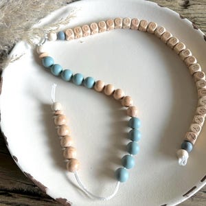 Może przedstawiać: Drewniany naszyjnik z koralikami alfabetu, koralikami silikonowymi w kolorze niebieskim i beżowym. Naszyjnik jest nawleczony na biały sznurek i ma drewnianą koralik z literą "A" na jednym końcu i drewnianą koralik z literą "Z" na drugim końcu.