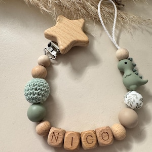 May include: A baby pacifier clip with wooden beads, a star-shaped wooden clip, and a green dinosaur charm. The clip features the name "NICO" spelled out in wooden letter blocks, and a crocheted bead.