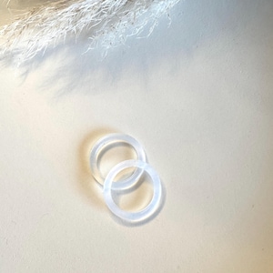 Könnte beinhalten: Zwei transparente, kreisförmige Ringe liegen auf einer hellbeigen Oberfläche. Die Ringe bestehen aus einem durchscheinenden Material und überlappen sich leicht. Der Hintergrund zeigt getrocknete, federartige Pflanzen.