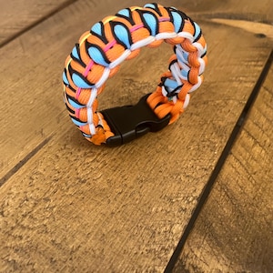 Könnte beinhalten: Ein farbenfrohes Paracord-Armband mit orangefarbenen, blauen, rosa und weißen gewebten Strängen. Es hat einen schwarzen Kunststoff-Schnallenverschluss. Das Armband wird auf einer Holzoberfläche präsentiert.