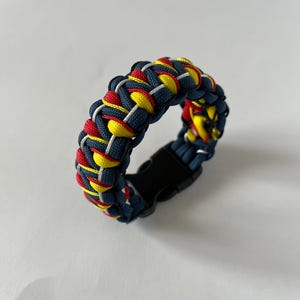 Könnte beinhalten: Geflochtenes Paracord-Armband in Marineblau, Rot, Gelb und Weiß. Das Armband hat einen schwarzen Kunststoff-Schnallenverschluss. Das gewebte Design erzeugt ein strukturiertes Aussehen, geeignet für Outdoor-Aktivitäten oder den Alltag.