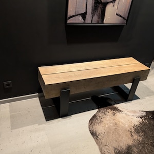 Könnte beinhalten: Eine rustikale Holzbank mit naturbelassenem Holz und schwarzen Metallbeinen. Die Bank steht auf einem hellgrauen Fliesenboden mit einem Kuhfellteppich davor.