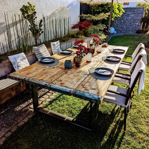 Könnte beinhalten: Ein rustikaler Holztisch mit Metallakzenten, gedeckt für ein Abendessen im Freien in einem Garten. Der Tisch ist von Stühlen umgeben und verfügt über ein Blumengebinde.
