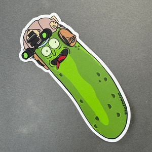 May include: A green pickle wearing a tan helmet with a green night vision scope and a brown pouch with the word "SPACE" on it. The pickle has a wide open mouth with a red tongue sticking out. The pickle has two large black eyes and a few black dots on its body. The sticker has the text "STALEFACE" on the bottom right corner.