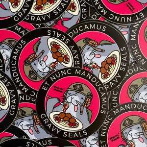 May include: A close-up of several round stickers featuring a cartoon seal wearing a military helmet and eating spaghetti and meatballs. The stickers are black and white with a pink background and the text "GRAVY SEALS ET NUNC MANDUCAMUS" in a white circle.