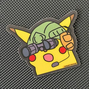 Può includere: Un adesivo di Pikachu giallo con un elmetto verde, cuffie arancioni e occhiali neri. L'adesivo ha guance rosse e bocca rosa. Il testo "STRIKEFORCE69" è stampato in basso a destra.