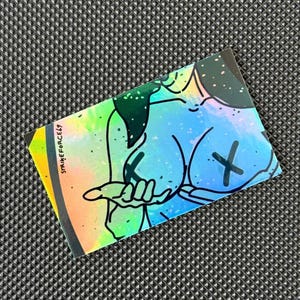 Può includere: Un adesivo olografico con un'illustrazione colorata di una figura con un segno X. L'adesivo ha una lucentezza arcobaleno e il testo "STRIKEFORCE69" è stampato sul lato sinistro.