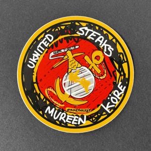 May include: A circular sticker featuring a red background with a yellow border and black scribbled accents. The text reads UKNITED STEAKS MUREEN KÖRE and STRIKEFORCE69. A central graphic depicts a gold eagle, globe, and anchor emblem with a banner.