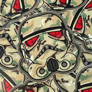 May include: A sticker featuring a stylised stormtrooper helmet with a camouflage pattern in shades of tan, green, and brown. The helmet has prominent red lenses for the eyes and black detailing around the mouth and side vents, outlined in a thick dark border.