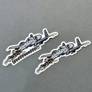 May include: Two white stickers featuring a cartoon illustration of a grey A-10 Warthog aircraft with a yellow and orange flame emanating from the engine. The text "BRRRRRRT!" is printed on the sticker.